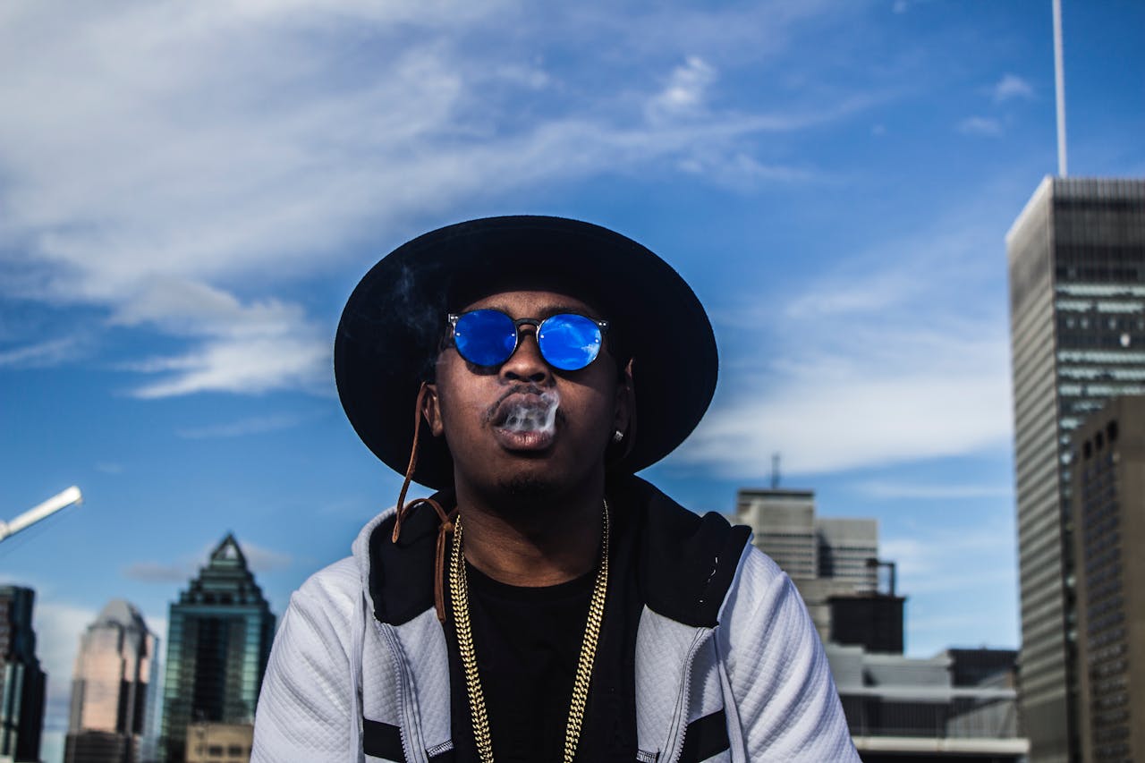 Stylish man in streetwear smokes against a cityscape backdrop, exuding urban cool.